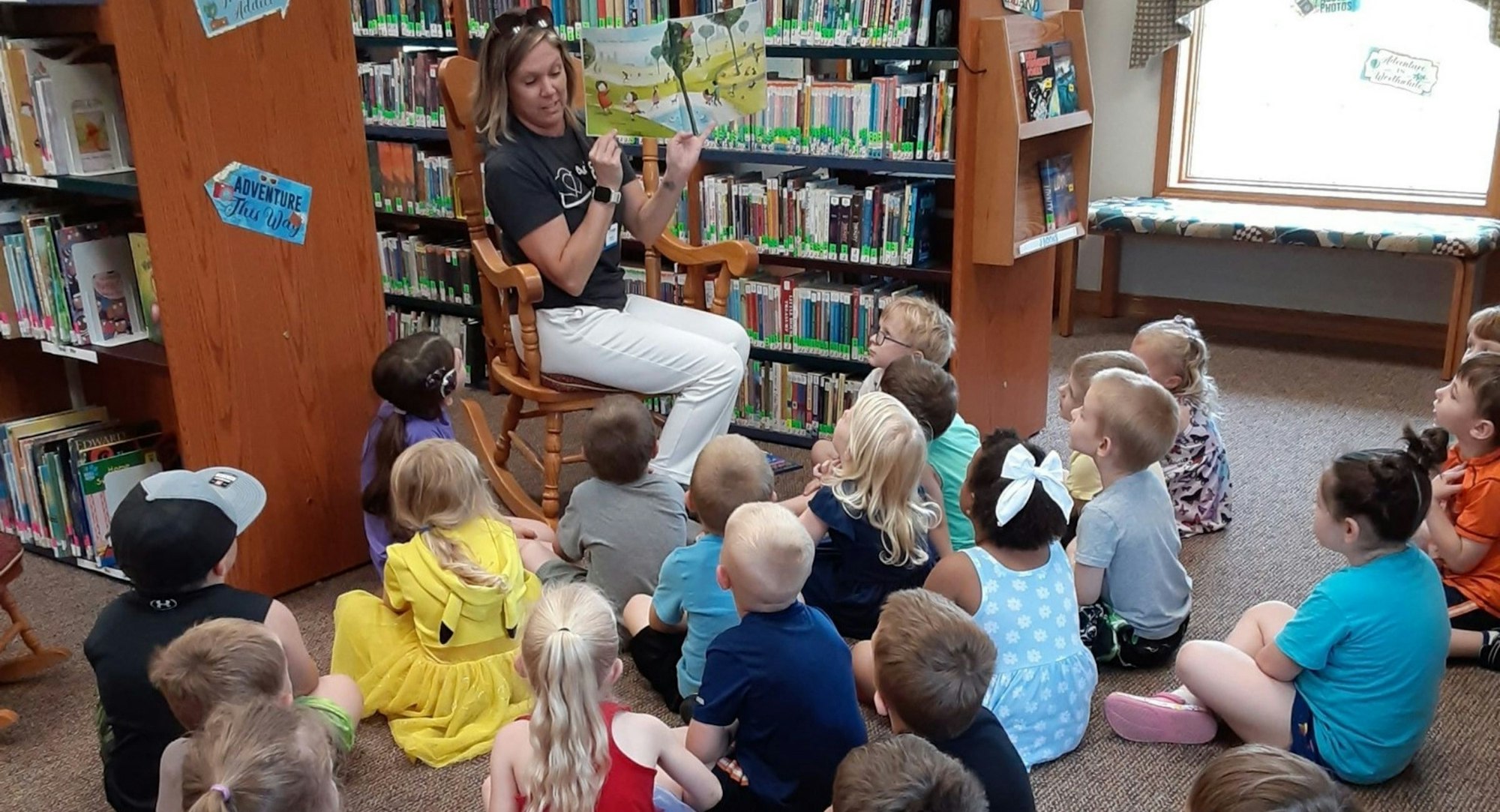A woman reads a story to a group of attentive kids in a library setting.