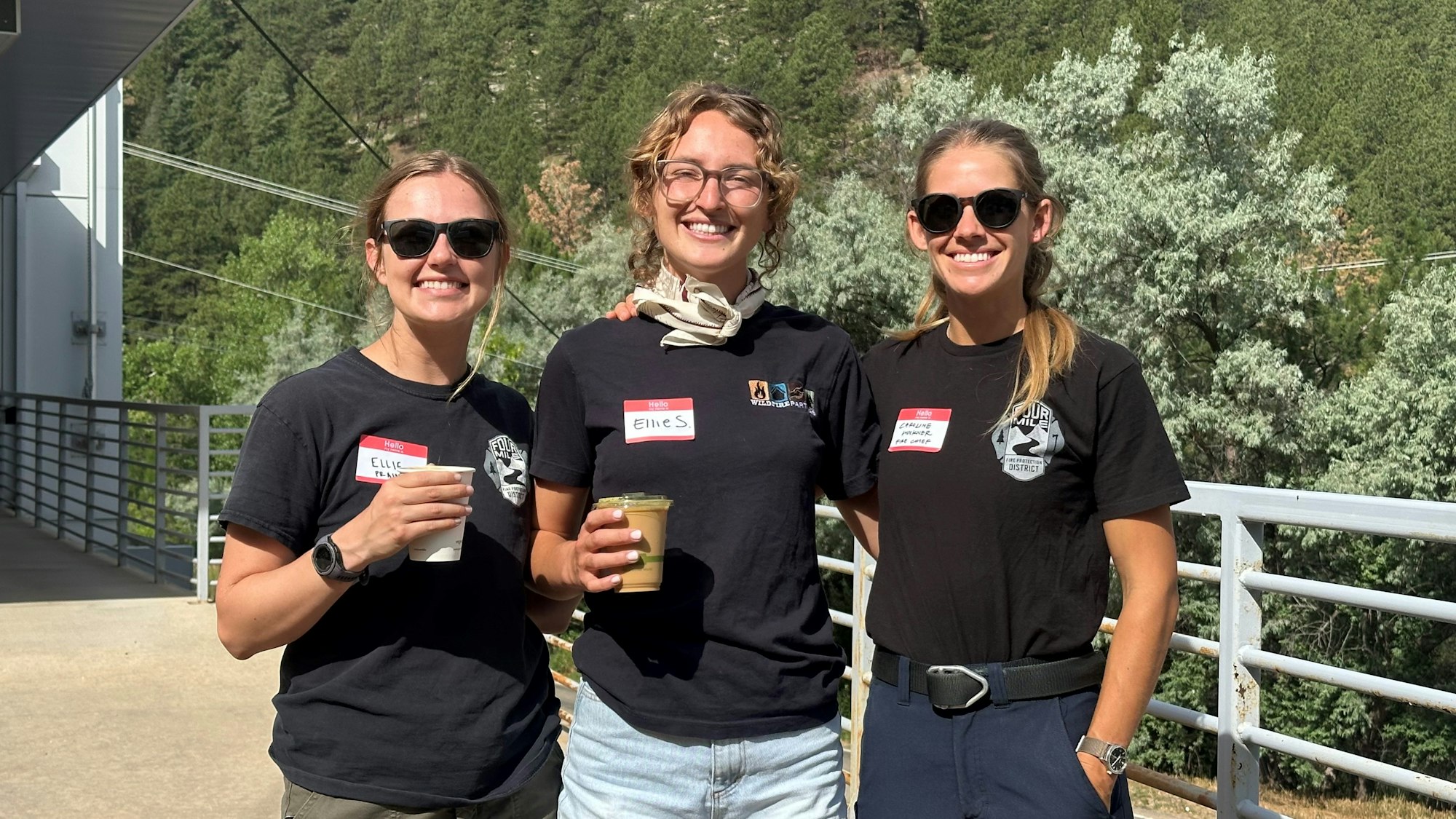 Ellie, Ellie and Chief Wockner with drinks, smiling outdoors with a forested background.