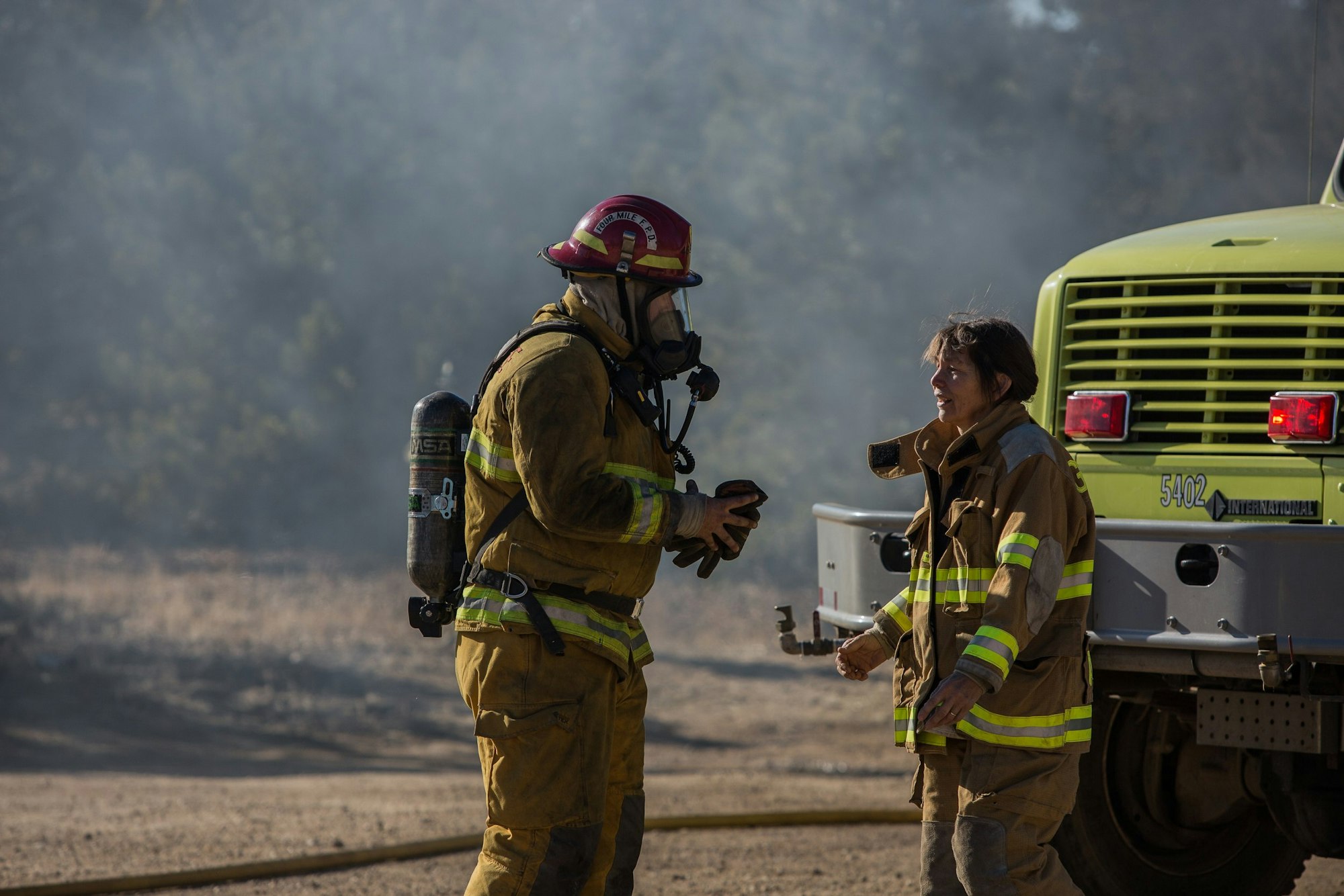 Two firefighters in protective gear near a fire truck, possibly discussing a wildfire response.