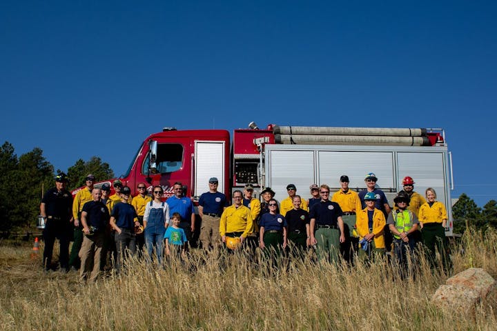 A group of firefighters in yellow and navy uniforms posing with a red fire engine in a grassy field.