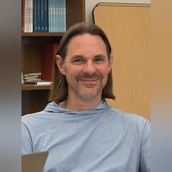 A man with long hair smiles, wearing a light blue hoodie, with shelves and books visible in the background.