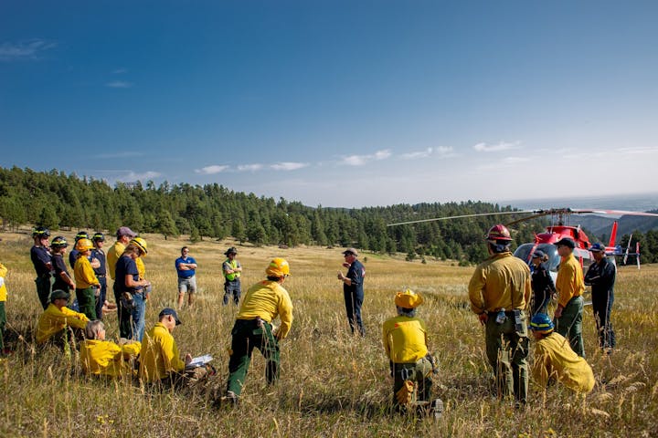 A group of firefighters in yellow gear gathering near a red helicopter in a grassy field.