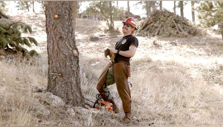 A person with a hard hat and sunglasses leaning on an axe, standing by a tree and a chainsaw, in a forest setting.