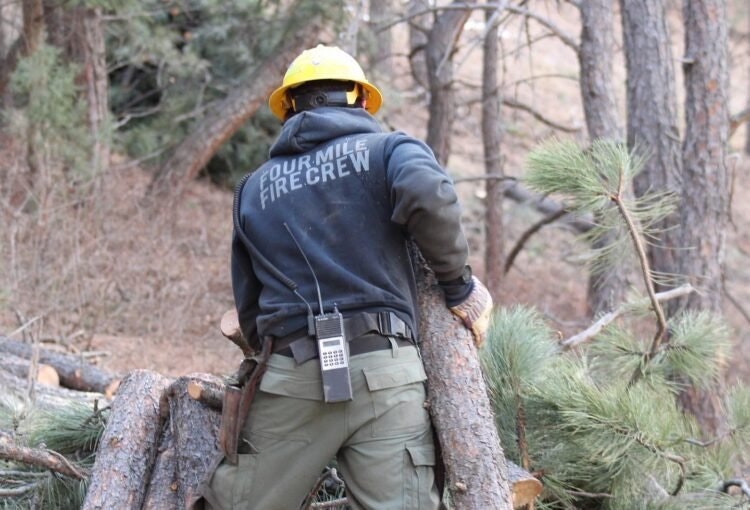 A firefighter in uniform marked "Fourmile Firecrew" with safety gear in a wooded area.