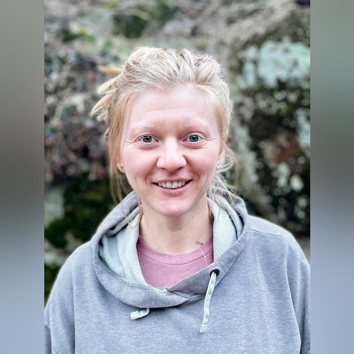 A smiling woman with blond hair in a gray hoodie, with foliage in the background.