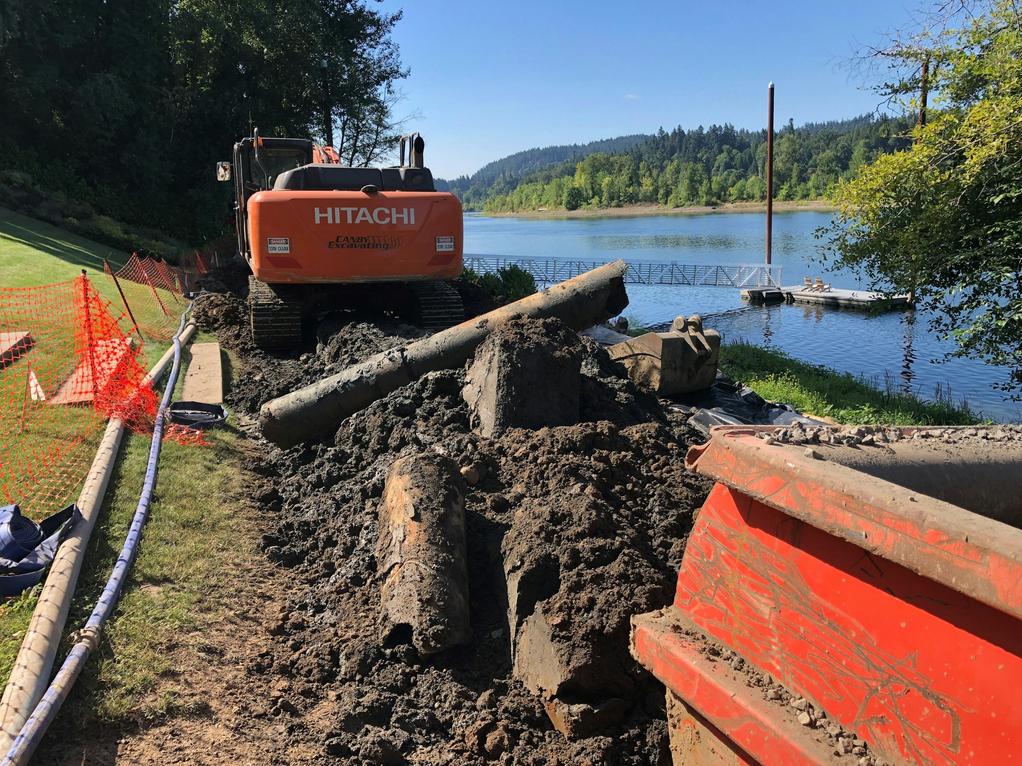 A construction site by a river featuring an excavator, dirt piles, and a visible dock in the background.