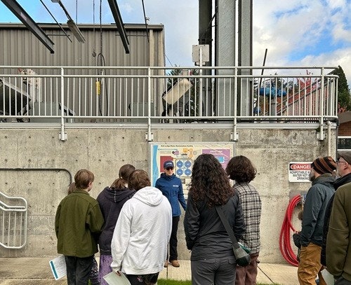 A group of people listen to a person speak in front of an industrial facility with safety signs and equipment.