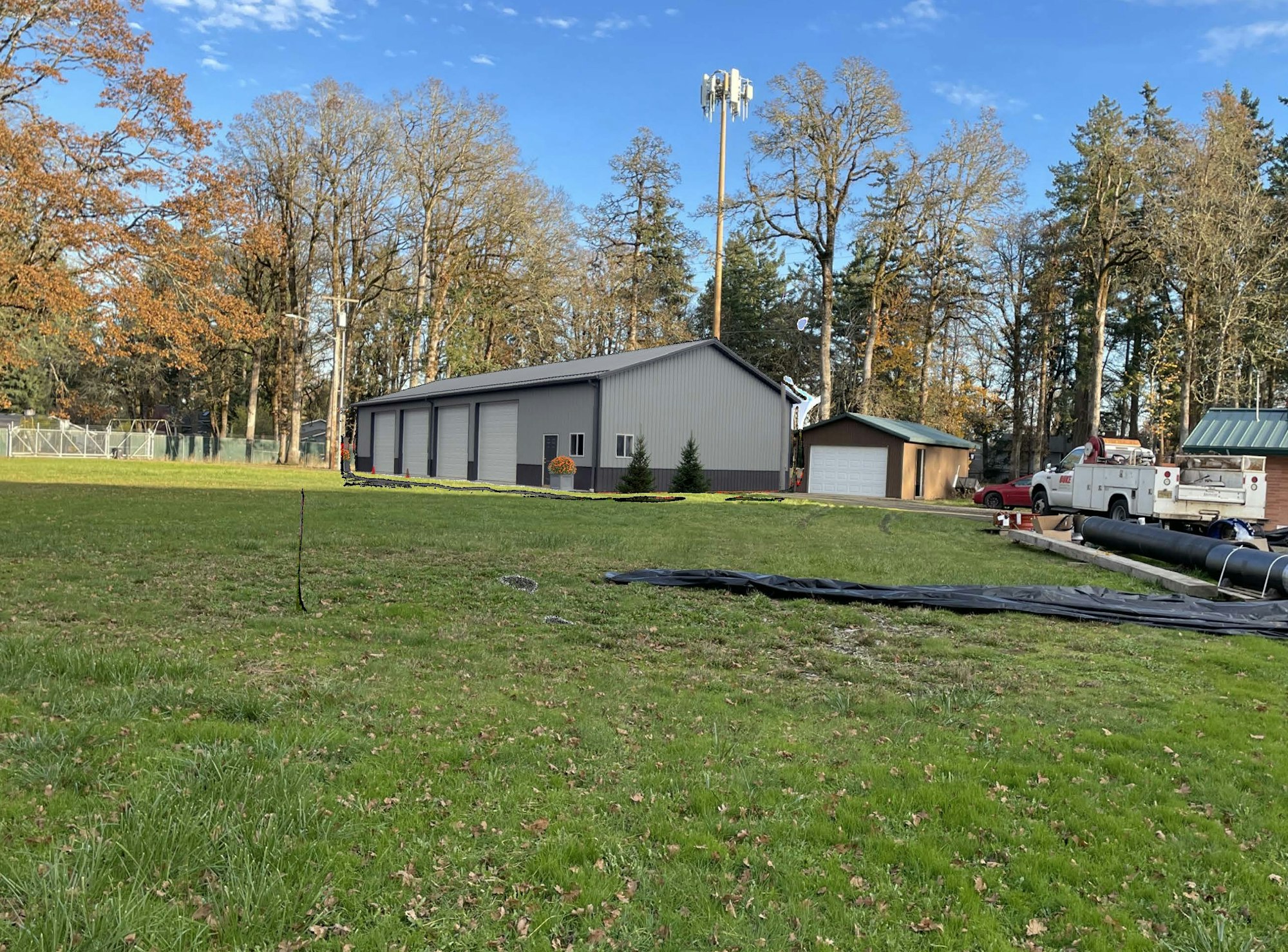 A metal building, utility trucks, and a grassy field with trees and a communication tower in the background.