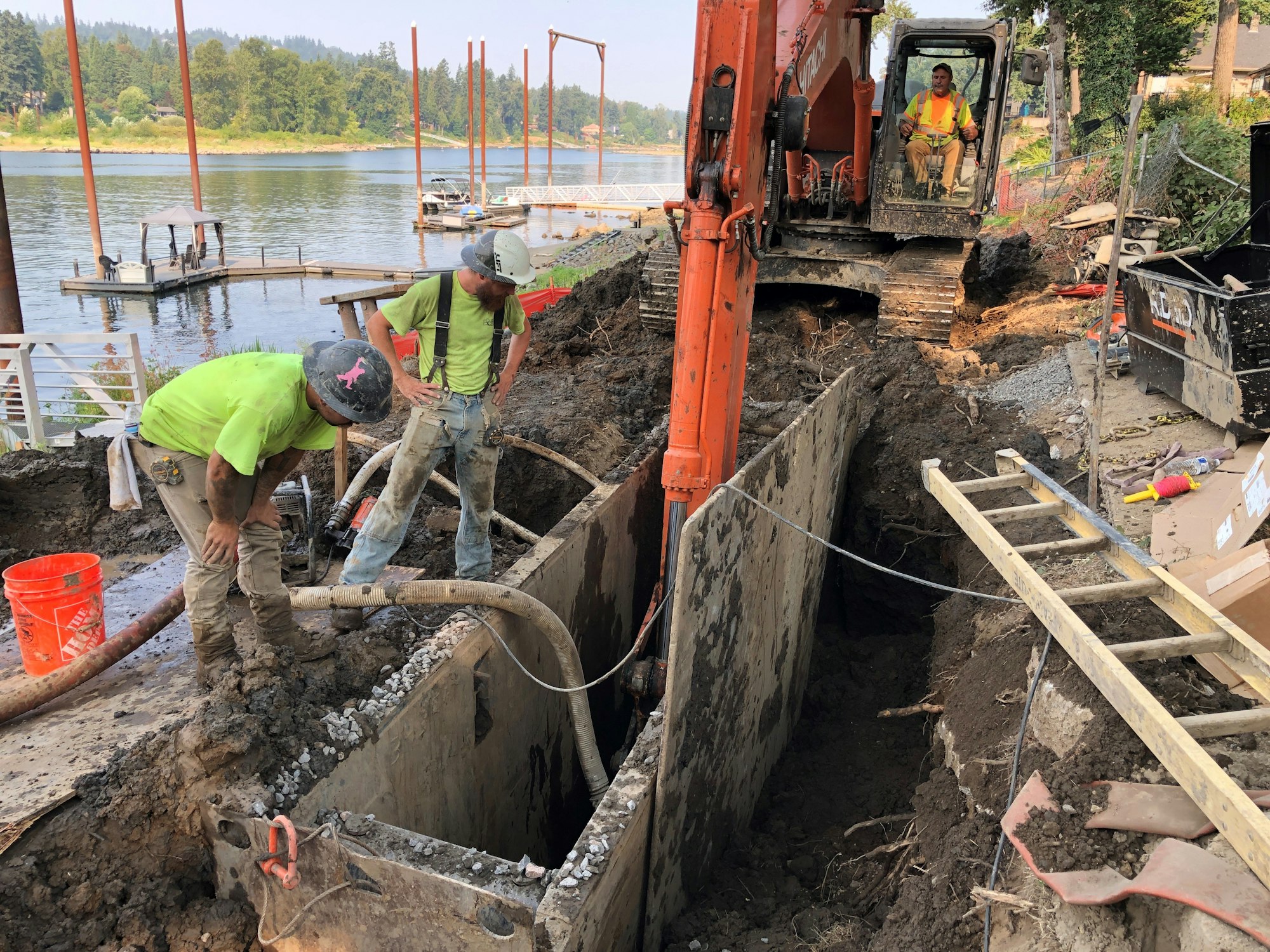 Construction workers are excavating near a river, using machines and tools to dig a large trench in the ground.