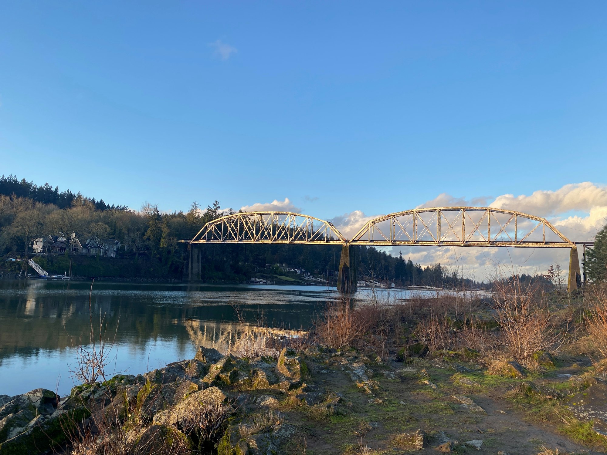 A truss bridge over a calm river with houses and trees, under a clear blue sky.