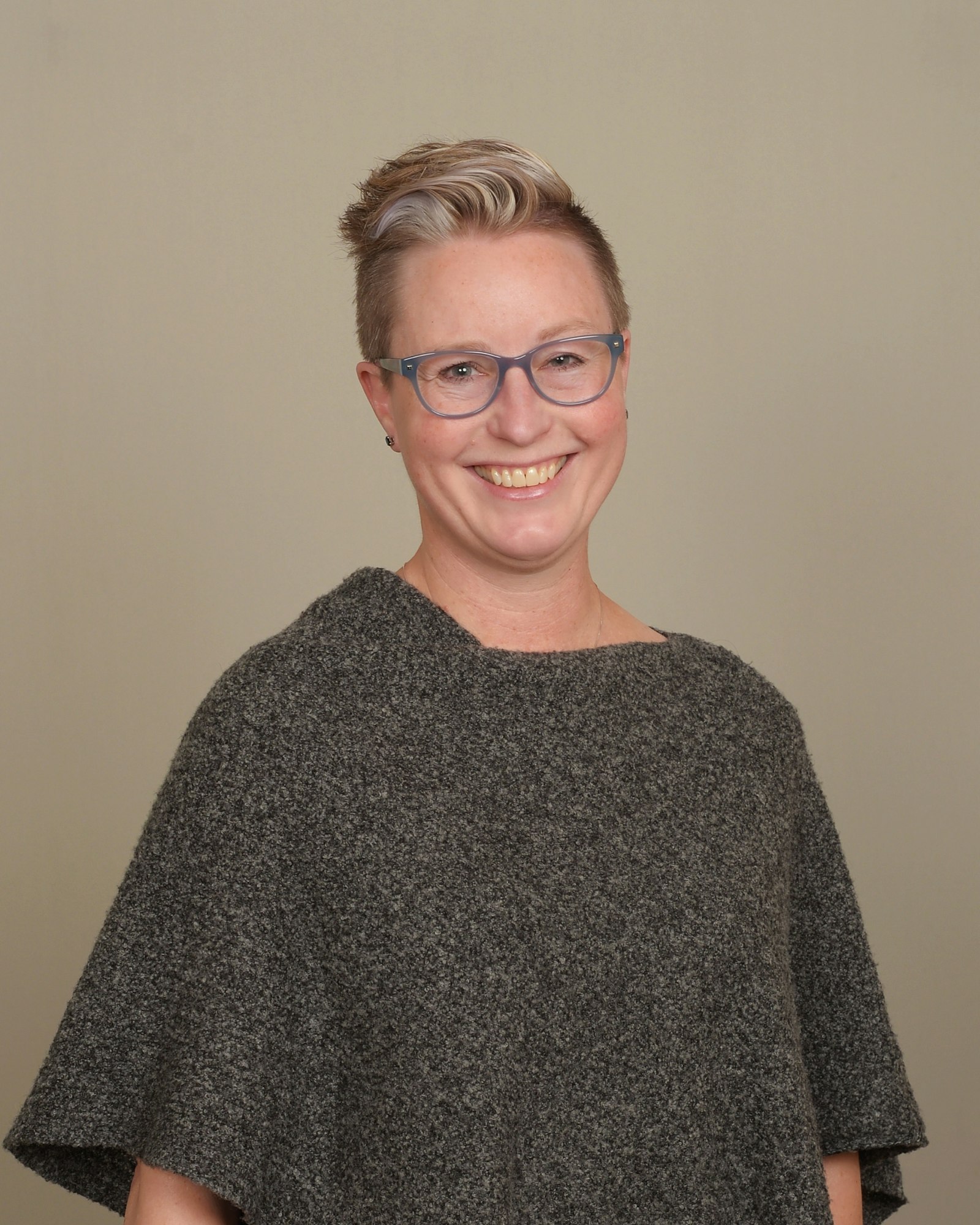 The image shows a smiling person with short hair, wearing glasses and a grey poncho against a neutral background.
