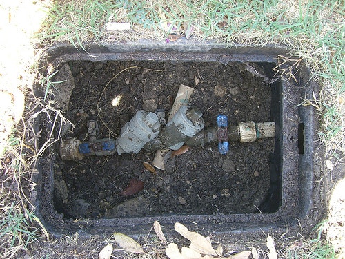 A buried irrigation system valve box, containing plumbing fixtures and dirt, surrounded by grass.