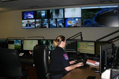 A person at a workstation with multiple monitors and screens displaying various data and maps on the wall.