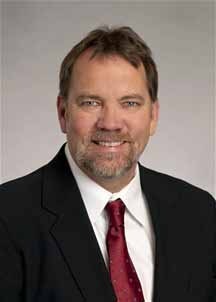 A man with a beard, wearing a black suit and red tie, smiling against a light background.