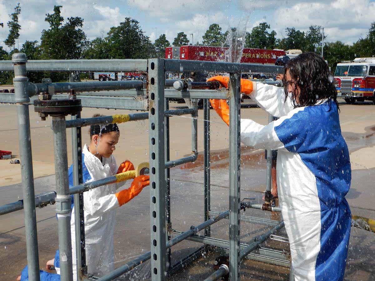 Two people in protective gear working with spraying water on a metal frame, possibly during an outdoor training exercise.