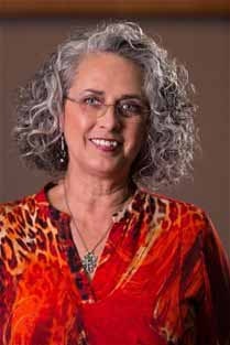 A smiling woman with curly gray hair, wearing glasses and a colorful blouse, with a pendant necklace.