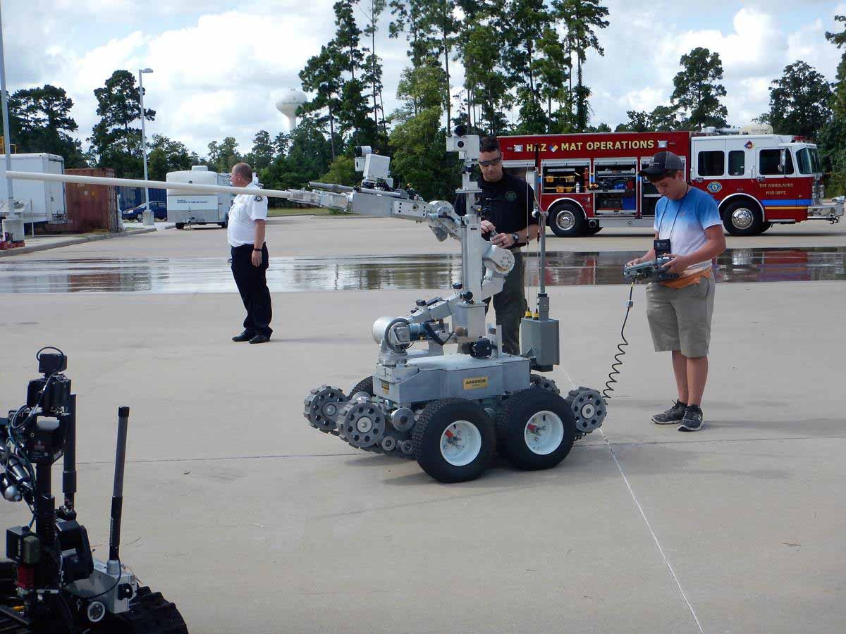 A bomb disposal robot being operated by technicians with a fire truck and a responder in the background.