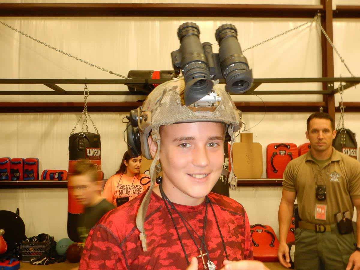 Person wearing helmet with large binoculars, in a room with boxing equipment and people in the background.