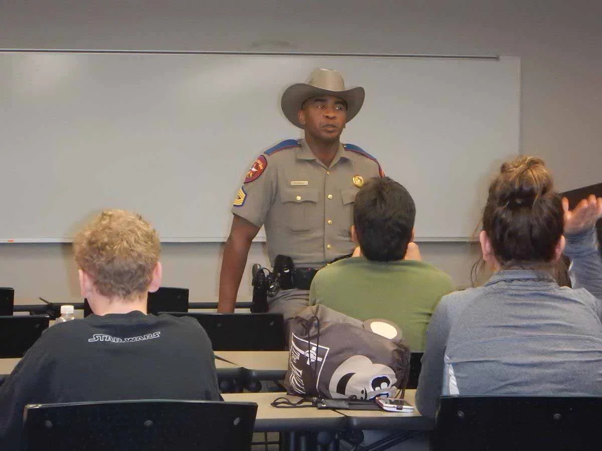 A uniformed officer stands speaking in a classroom with students listening.