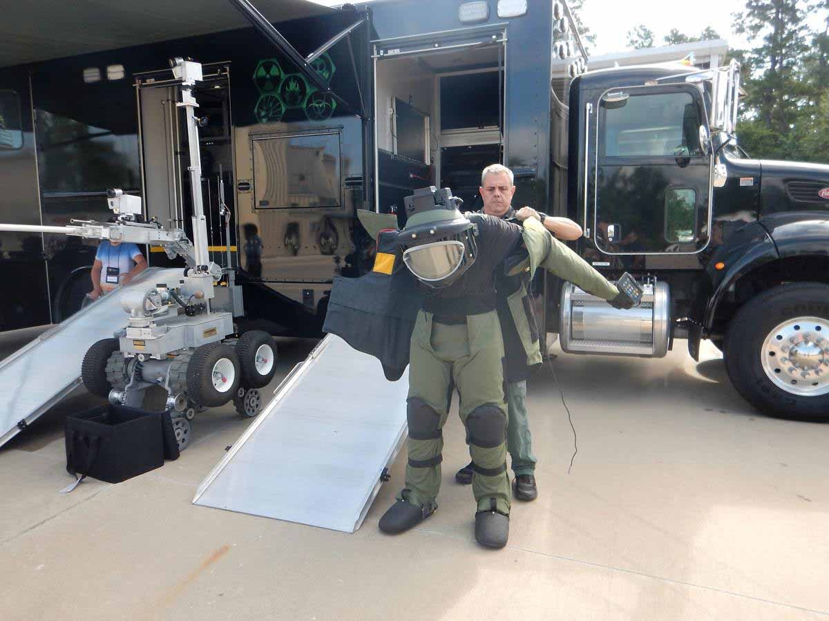 Man in bomb disposal suit beside a robot and specialized vehicle.