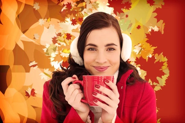 A smiling person wearing earmuffs holds a red mug, with a backdrop of autumn leaves.