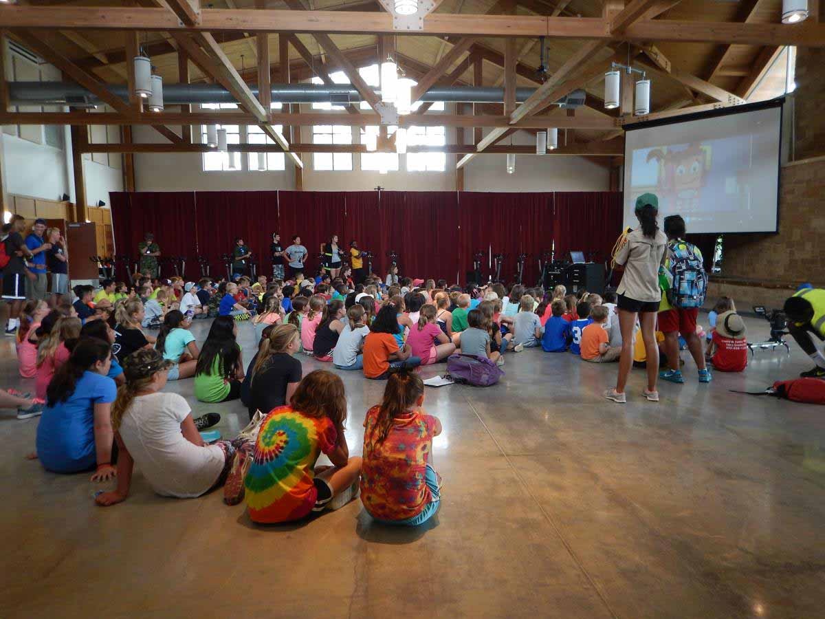 Children sitting on the floor in a hall, watching a presentation with a cartoon on screen, while adults supervise.