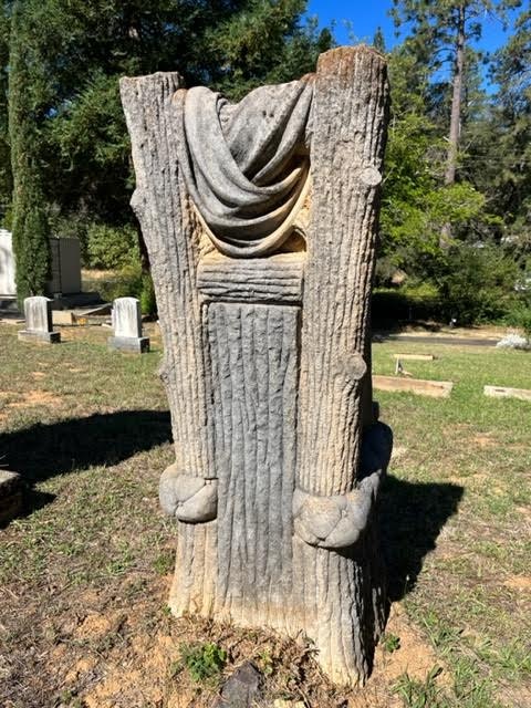 A sculpted tree stump grave marker with draped fabric detail, in a cemetery setting.