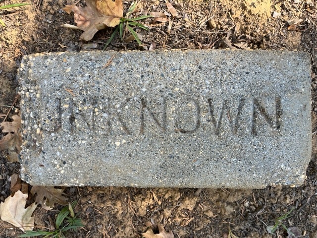 A weathered stone engraved with "UNKNOWN" lying on the ground among dry leaves.