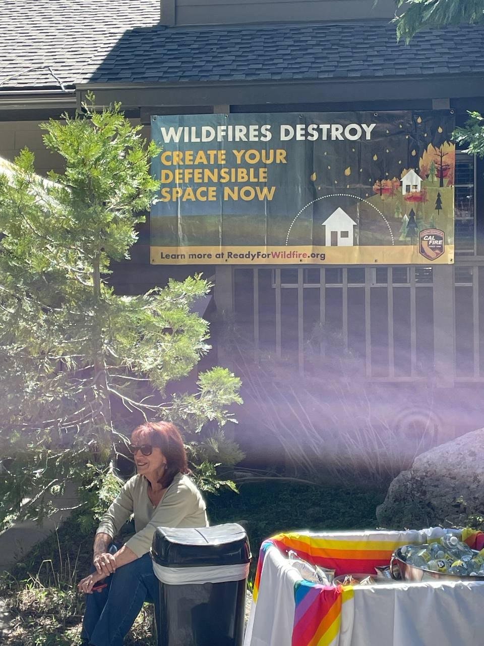 A woman sits beside a trash can, with a banner urging wildfire safety and creating defensible space, in a community setting.