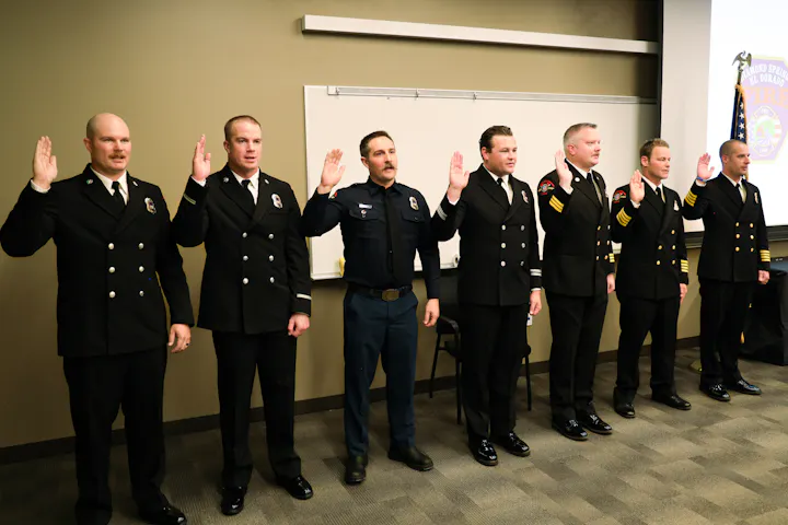 Firefighters are taking an oath, standing in uniform with raised hands, in a formal setting.