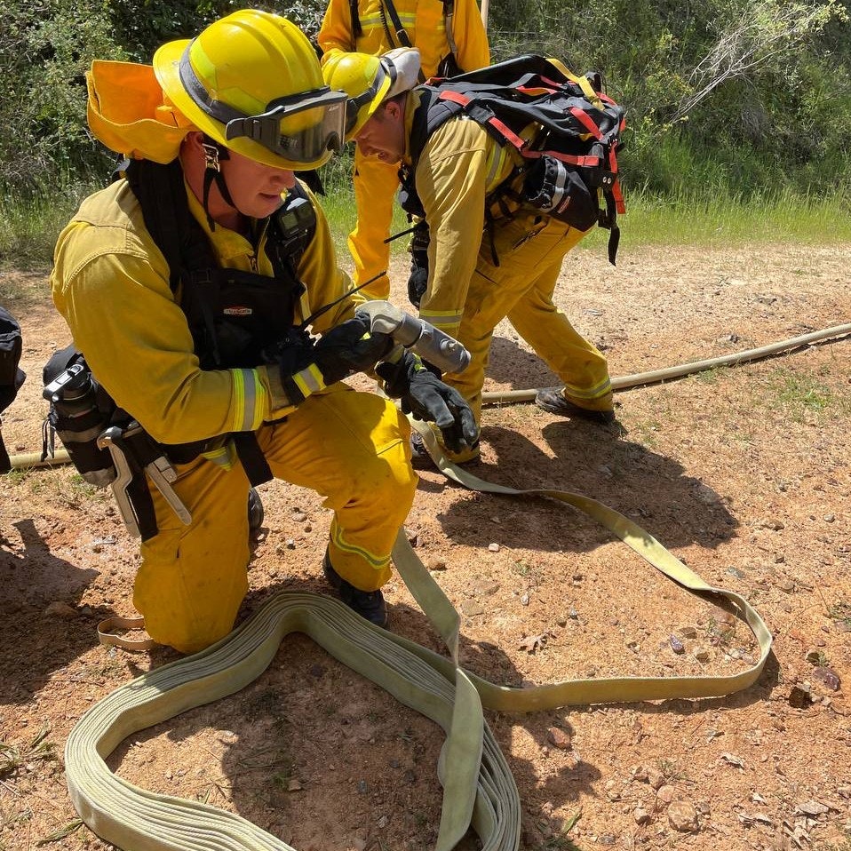 Firefighters in yellow gear are working on setting up equipment in a wilderness area, focusing on a hose and gear.