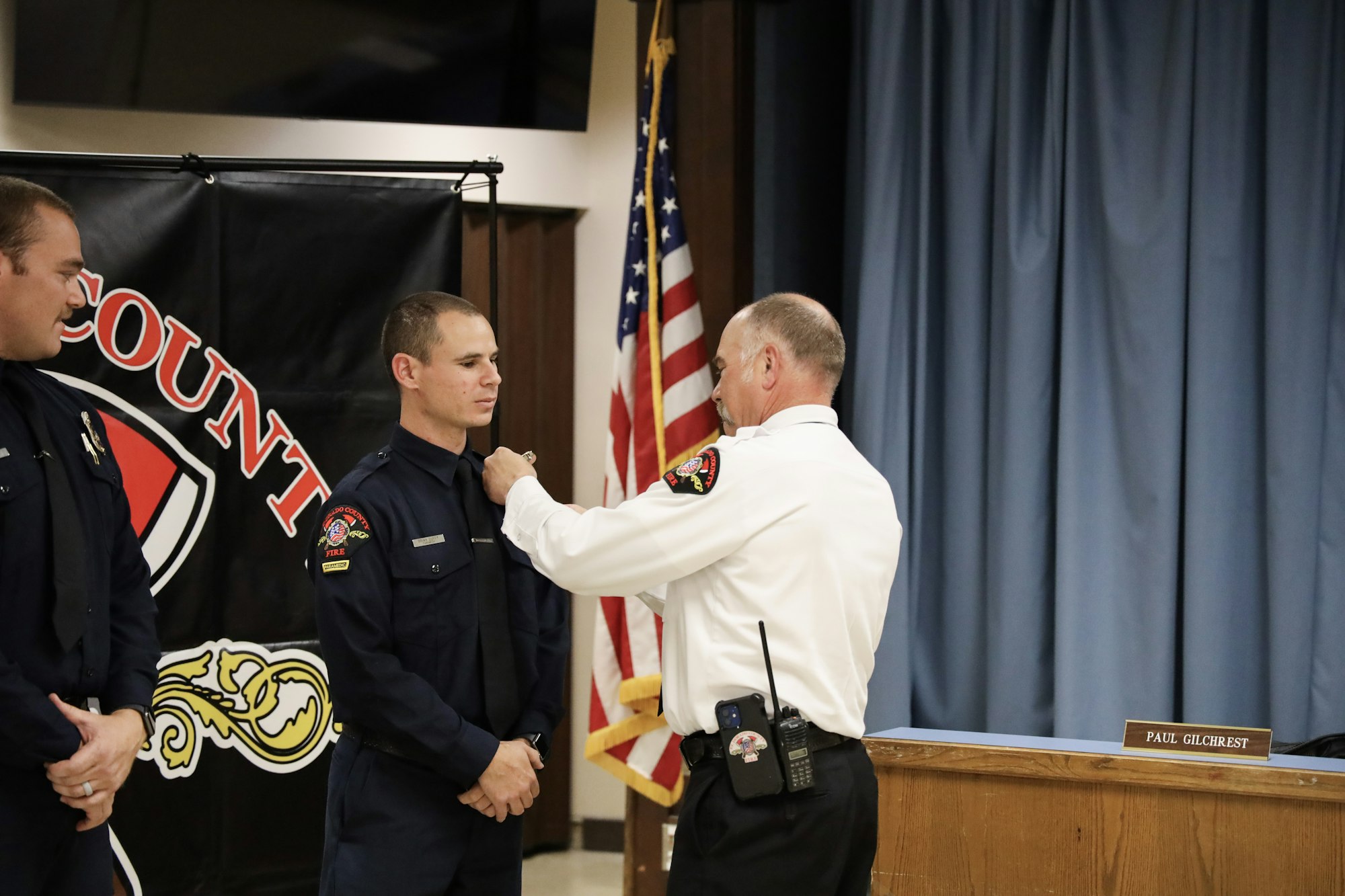 A ceremony with a fire department official pinning a badge onto the uniform of a firefighter, with an American flag backdrop.