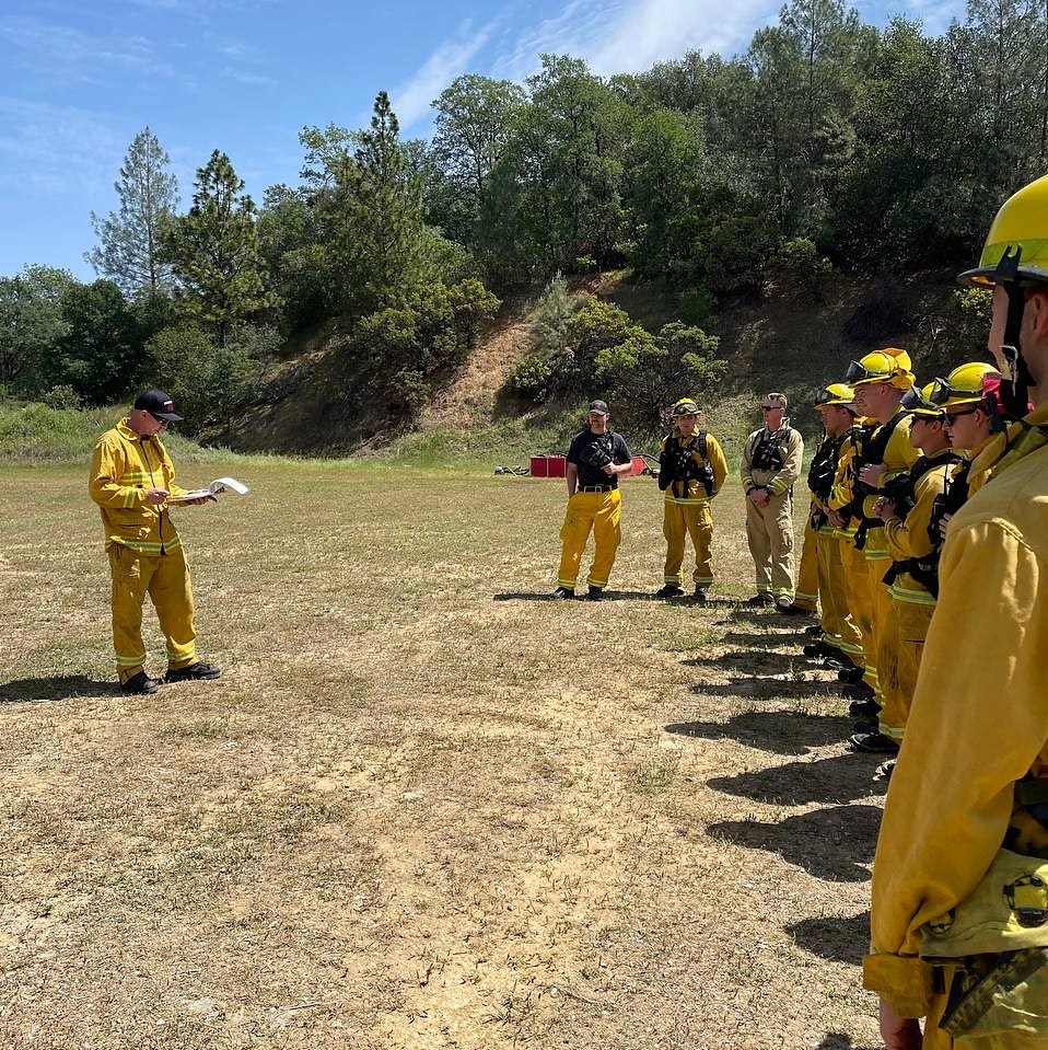 A group of firefighters in yellow gear listens to a leader during a training session in a sunny outdoor setting.