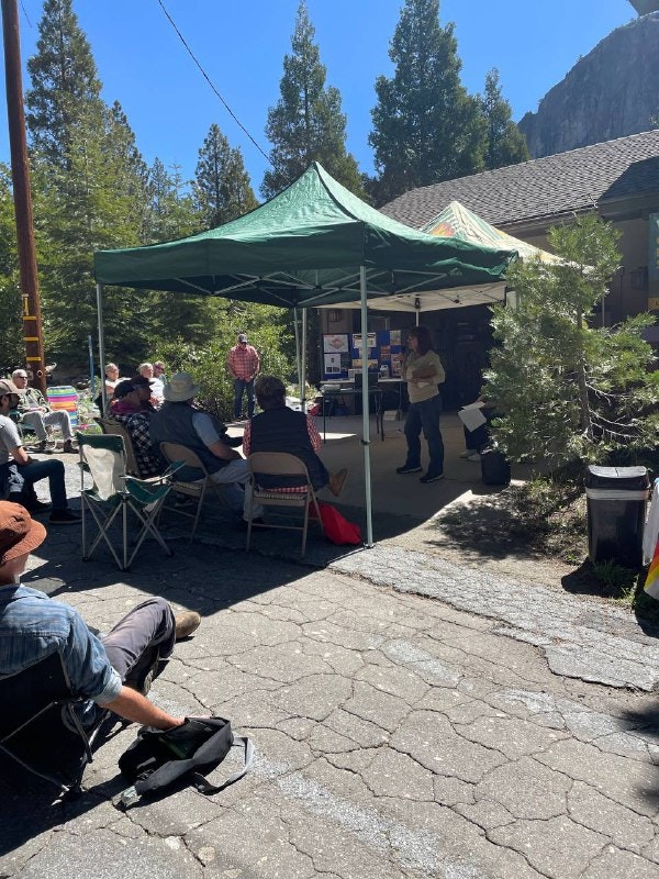A group gathers under a tent for a presentation outdoors, surrounded by trees and a mountainous backdrop.