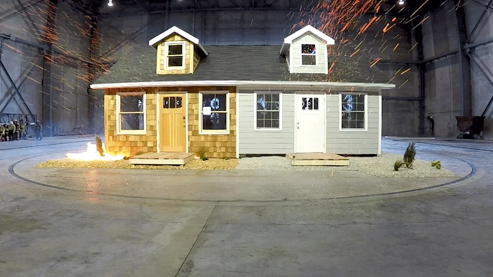 animation of a small test house with flying embers. one side made of exposed wood and flammable landscape burns while a second half that is hardened from wildfire does not.