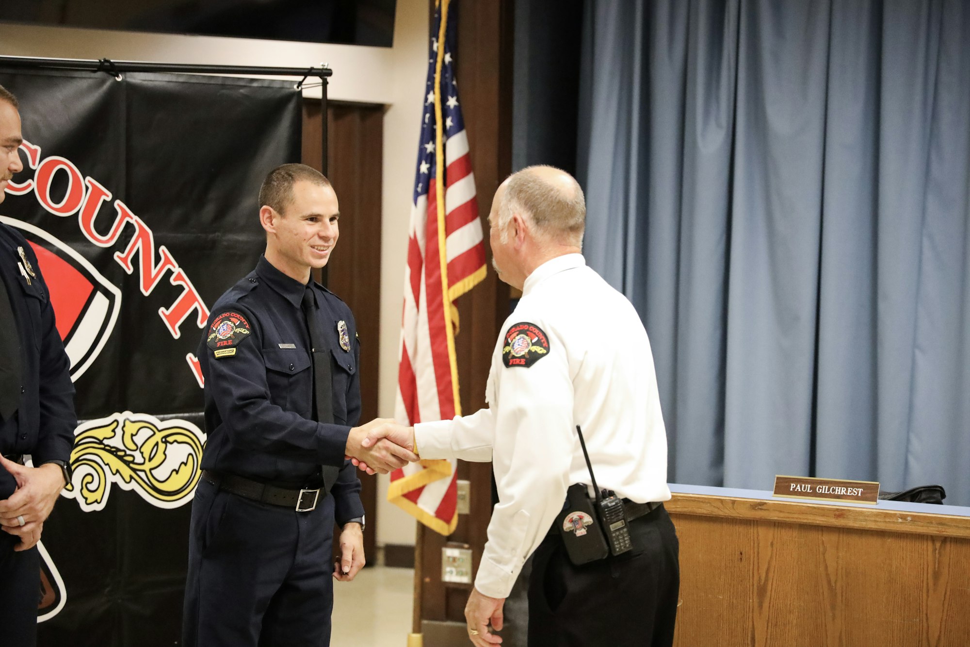 Firefighters in uniform shaking hands at a formal event with a US flag and insignia banner in the background.