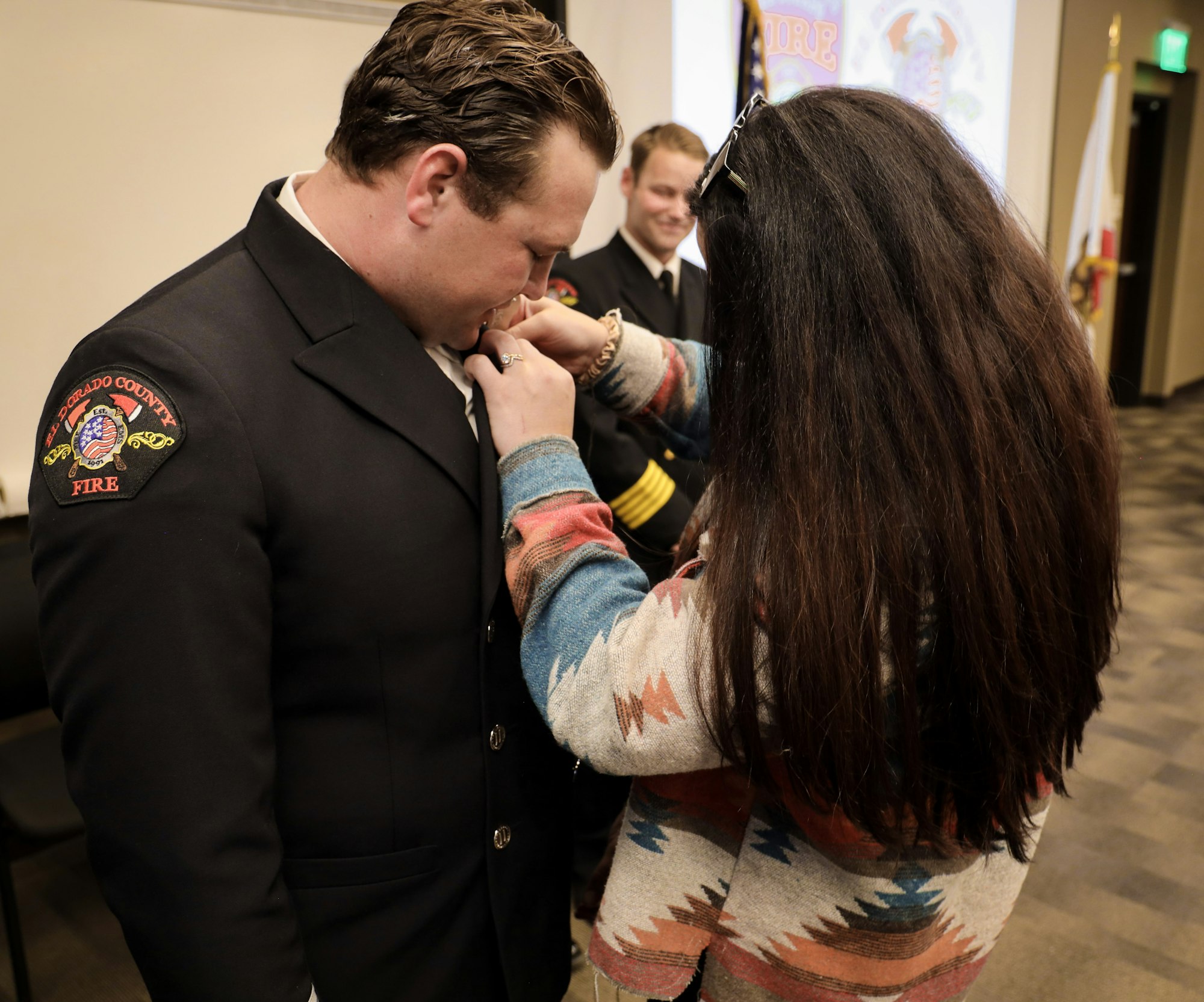 A woman is pinning a badge on a man in a fire department uniform during a ceremony, while another man looks on.