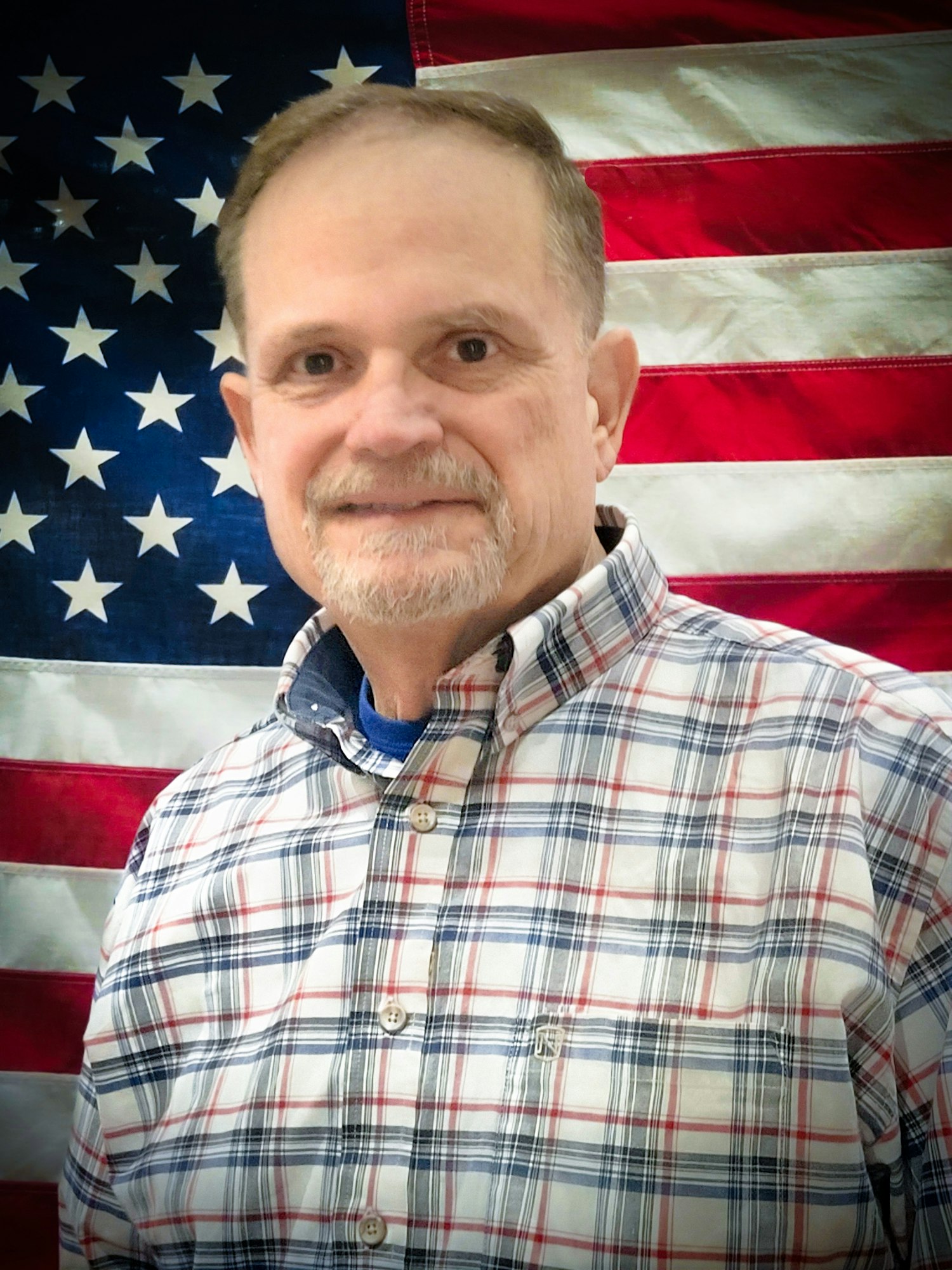 A man in a plaid shirt standing in front of an American flag.