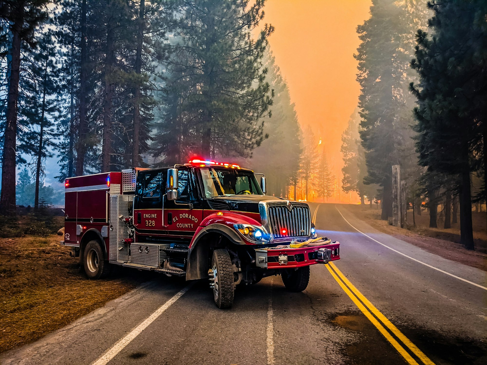 A fire engine is parked on a smoky road, surrounded by trees and an approaching wildfire in the background.