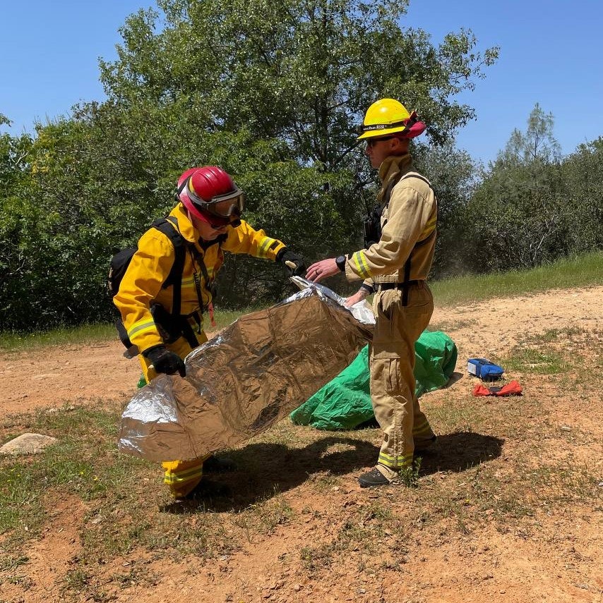 Two rescue workers are seen assisting with a reflective thermal blanket outdoors, likely during a rescue operation.