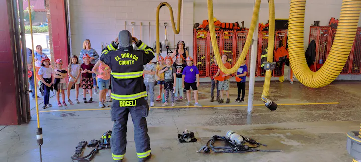 A firefighter is demonstrating safety to a group of kids wearing helmets in a fire station. Equipment is also visible on the floor.