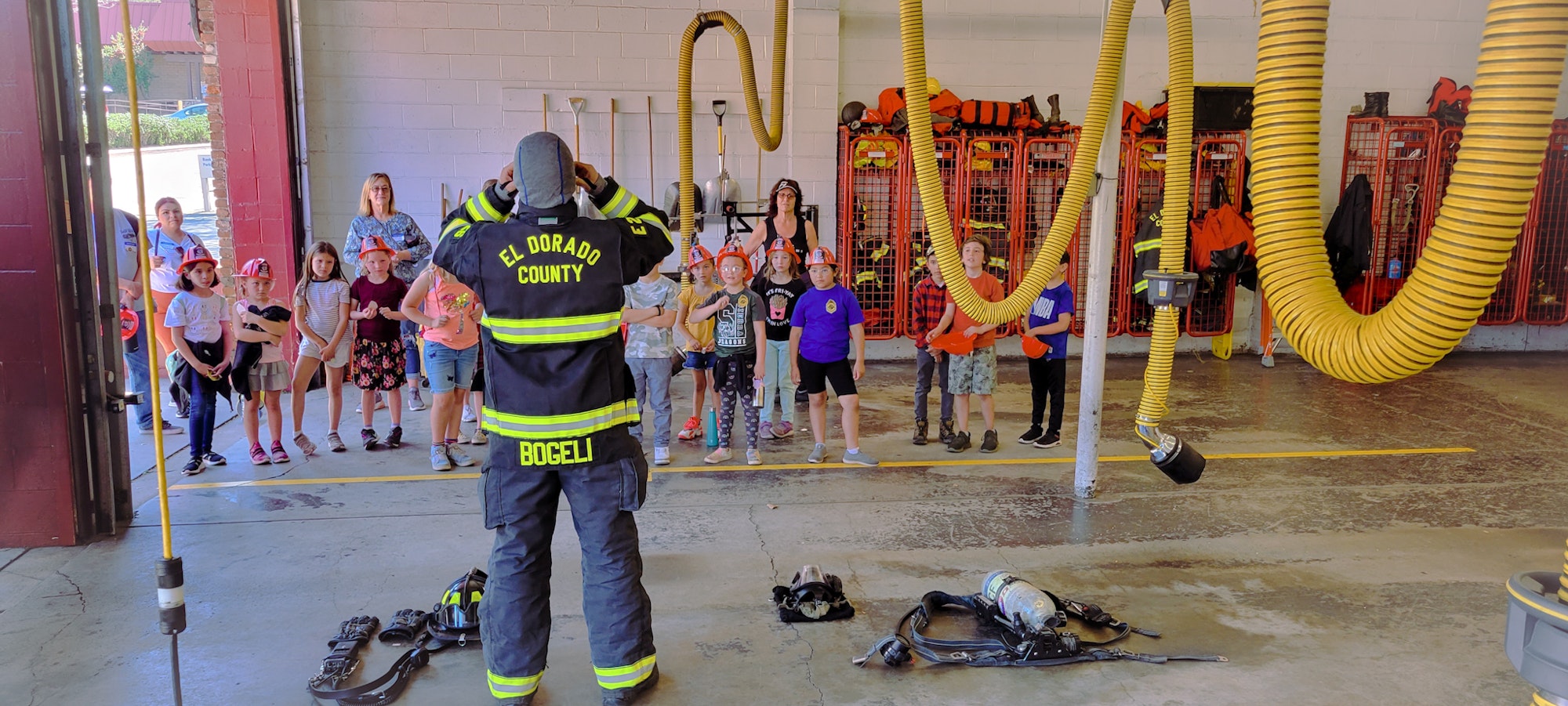 A firefighter is demonstrating safety to a group of kids wearing helmets in a fire station. Equipment is also visible on the floor.