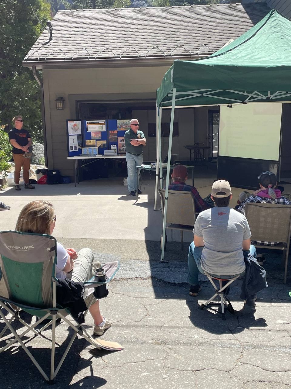 A presentation is taking place outdoors, with an audience seated and a speaker discussing topics near display boards.