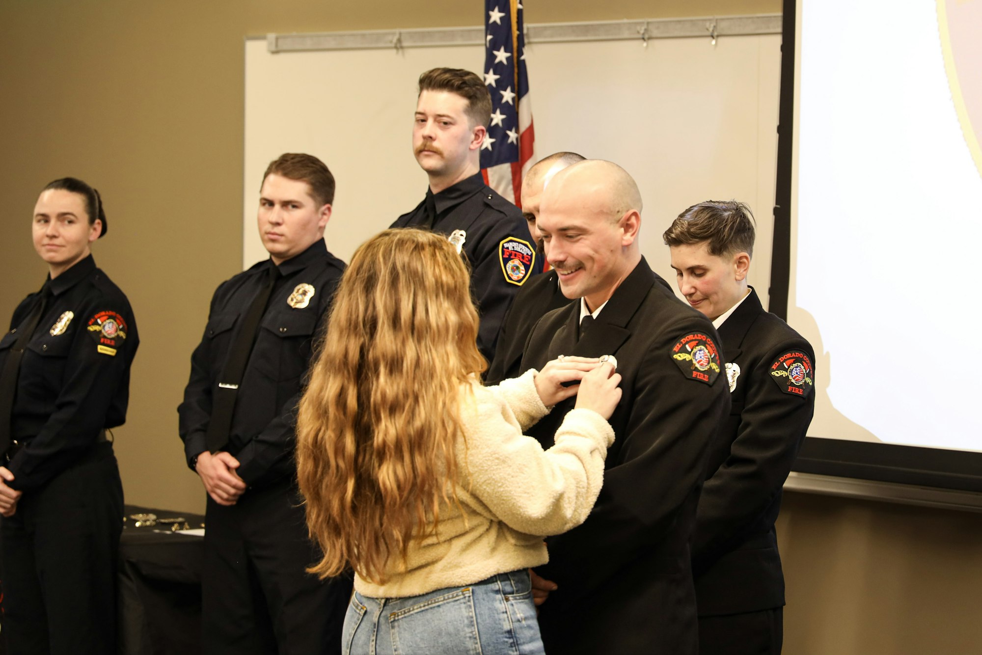 A woman pins a badge on a smiling firefighter in uniform, with others standing nearby.