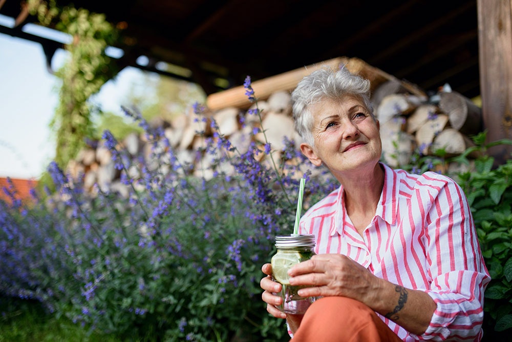 An elderly woman with a drink, smiling outdoors surrounded by plants.