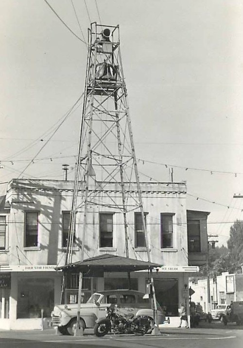 black and white photo containing streets, 1950's police car, motorcycle, bell tower, building in background.