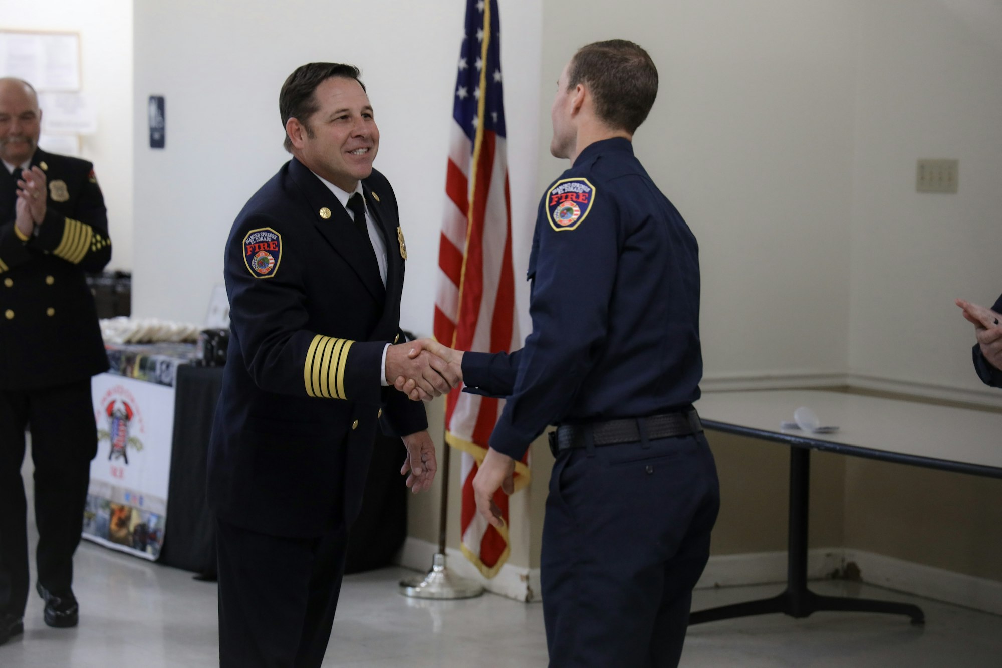 Two firefighters in uniform shaking hands, one with captain's bars, American flag in the background, clapping colleague nearby.