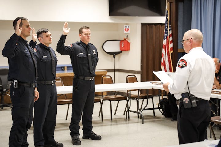 Three firefighters taking an oath led by an officer in a room with an American flag.