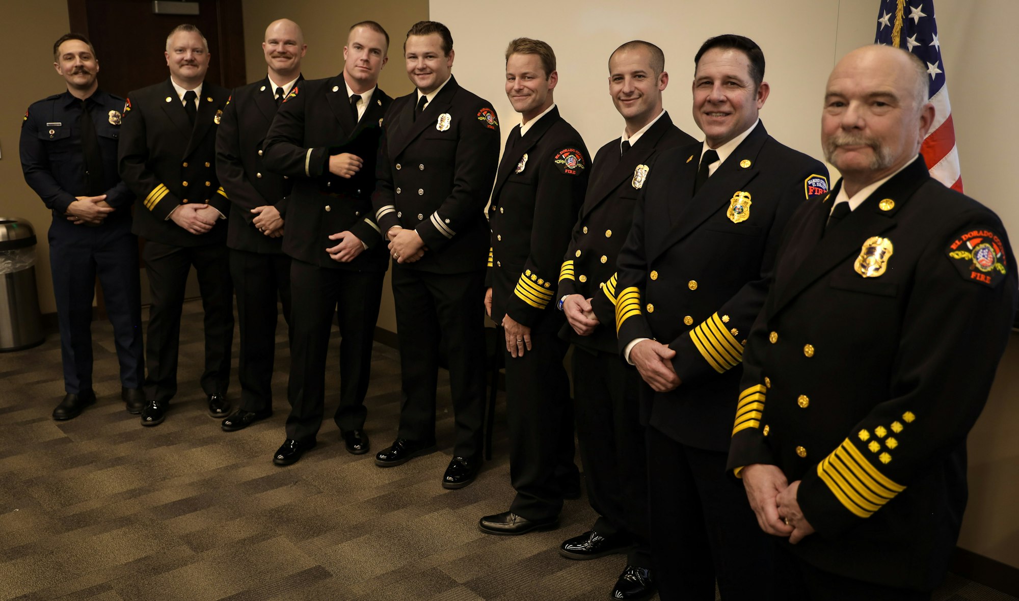 A group of firefighters in formal uniforms stands together in a room, with an American flag in the background.