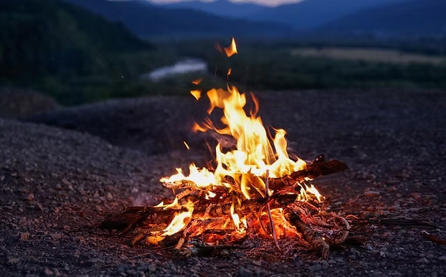 A campfire burning on a rocky surface with a scenic mountain and river landscape in the background.